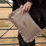 A person outdoors holds a brown and beige gingham clutch with ruffled edges and the word 'chloe embroidered on the front. The person is wearing a dark coat, black leggings, and black rain boots, with a dry field and metal gate in the background.