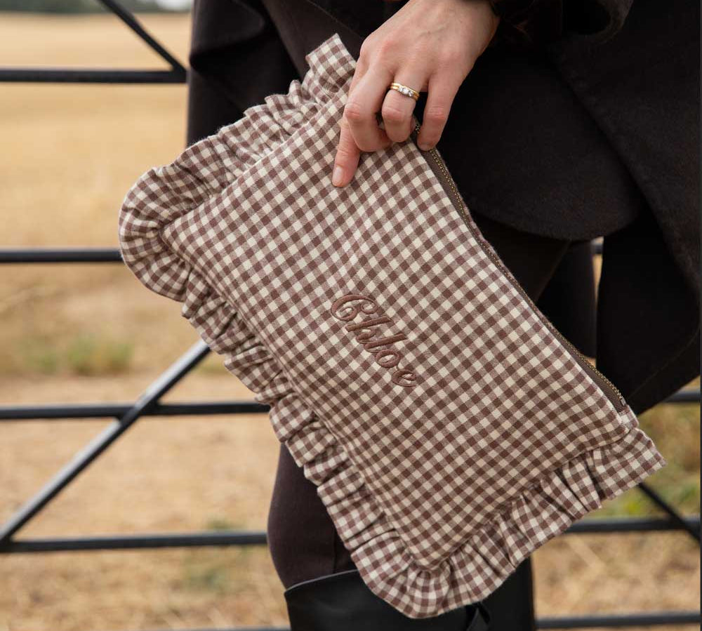 A person outdoors holds a brown and beige gingham clutch with ruffled edges and the word 'chloe embroidered on the front. The person is wearing a dark coat, black leggings, and black rain boots, with a dry field and metal gate in the background.