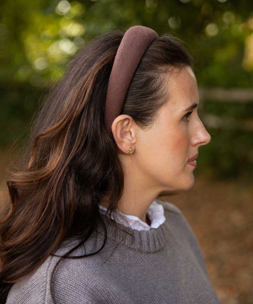 A woman in profile wearing a wide, brown headband and a gray knit sweater, photographed outdoors with a soft blurred green and brown background.