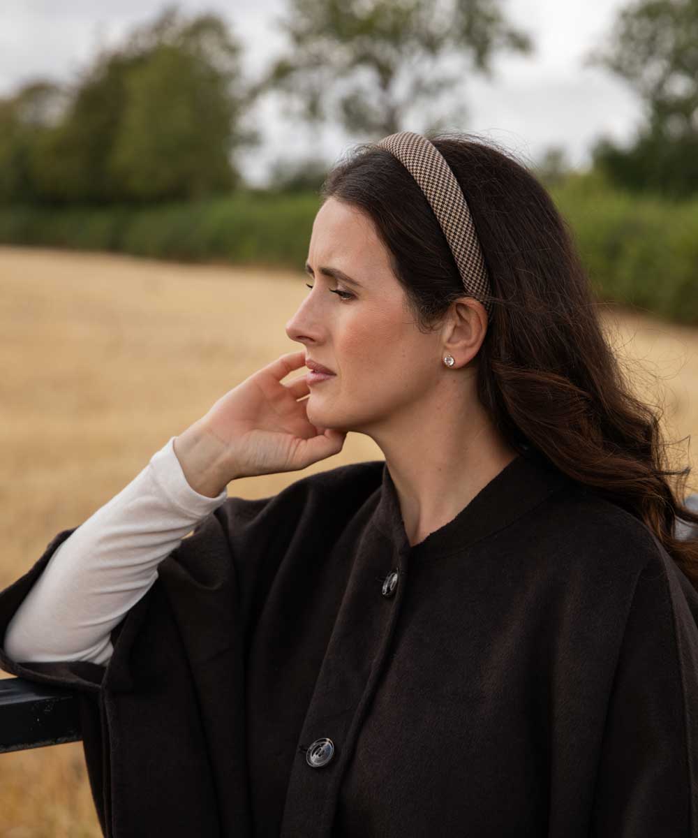 A woman in a black coat and white long-sleeve shirt wearing a brown patterned headband, posing outdoors in a field.