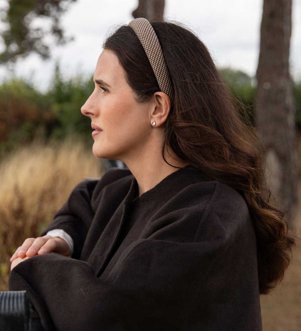 A woman in a dark brown wool coat and a beige patterned headband is shown in profile outdoors against a soft-focus background of trees and dry grass.