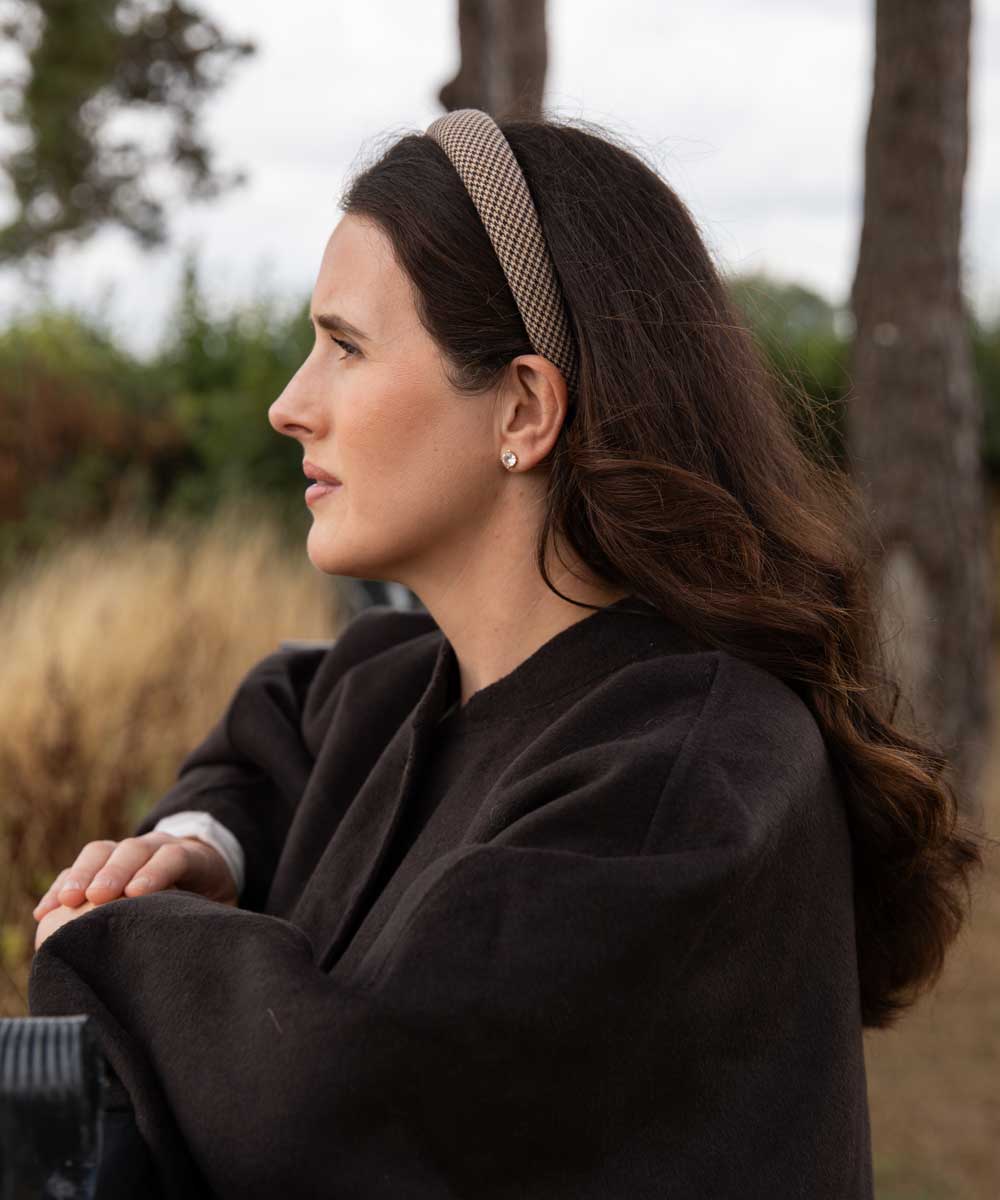 A woman in a dark brown wool coat and a beige patterned headband is shown in profile outdoors against a soft-focus background of trees and dry grass.