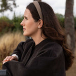 A woman in a dark brown wool coat and a beige patterned headband is shown in profile outdoors against a soft-focus background of trees and dry grass.