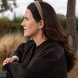 A woman in a dark brown wool coat and a beige patterned headband is shown in profile outdoors against a soft-focus background of trees and dry grass.