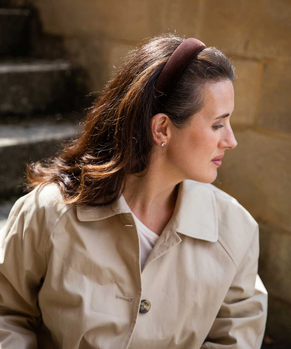 A woman in profile wearing a beige trench coat and a dark brown wide headband, set against a blurred stone wall background.