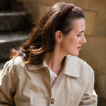 A woman in profile wearing a beige trench coat and a dark brown wide headband, set against a blurred stone wall background.