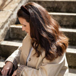 A woman with long brown hair sits on outdoor stone steps, wearing a beige trench coat over a white top and a checkered shirt, with a dark brown headband.