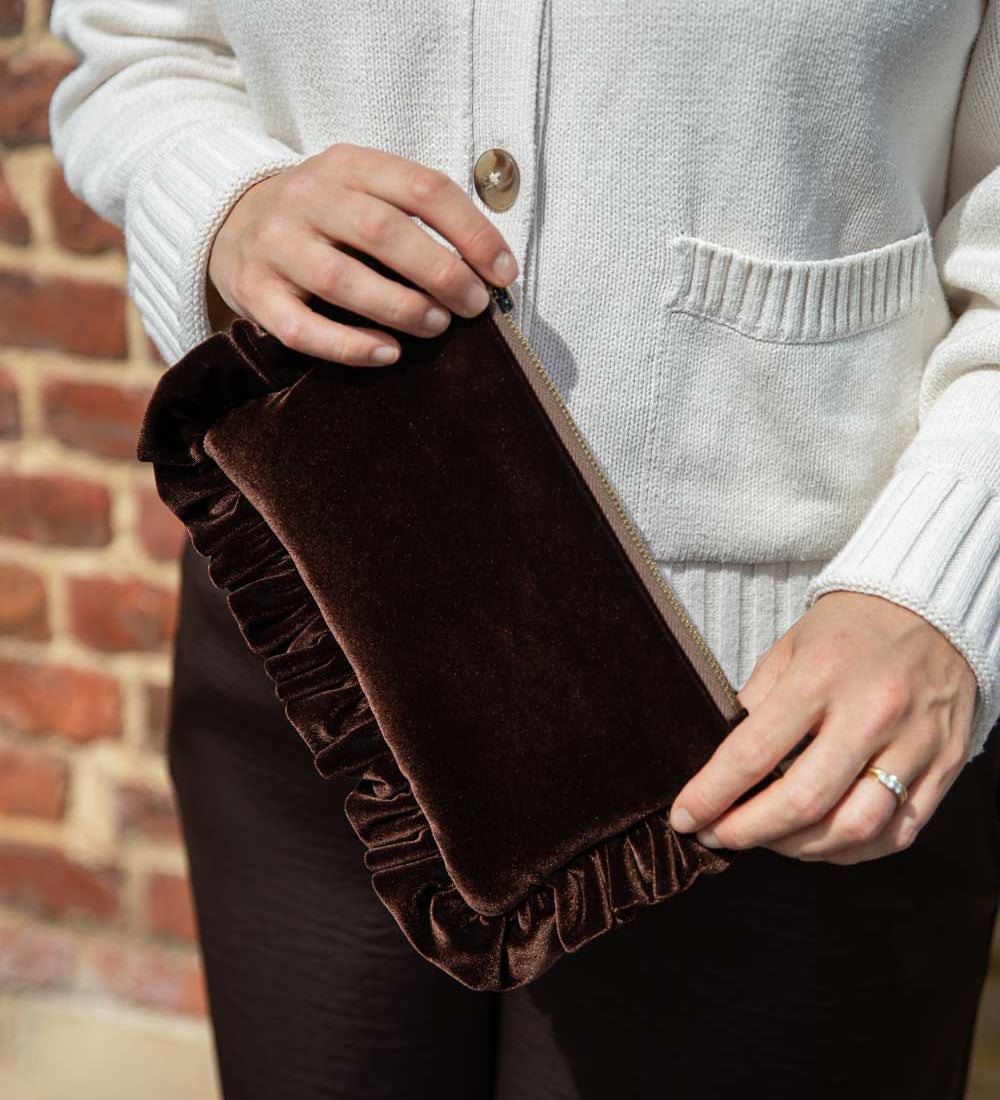 A person wearing a cream ribbed cardigan holds a dark brown velvet clutch bag with a ruffled edge and a beige zipper, set against a brick wall background.