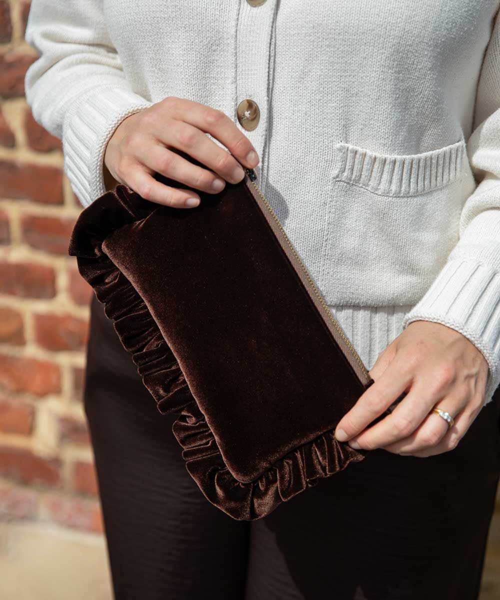 A person wearing a cream ribbed cardigan holds a dark brown velvet clutch bag with a ruffled edge and a beige zipper, set against a brick wall background.