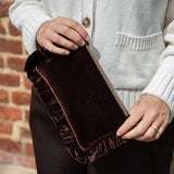 A person wearing a cream ribbed cardigan holds a dark brown velvet clutch bag with a ruffled edge and a beige zipper, set against a brick wall background.
