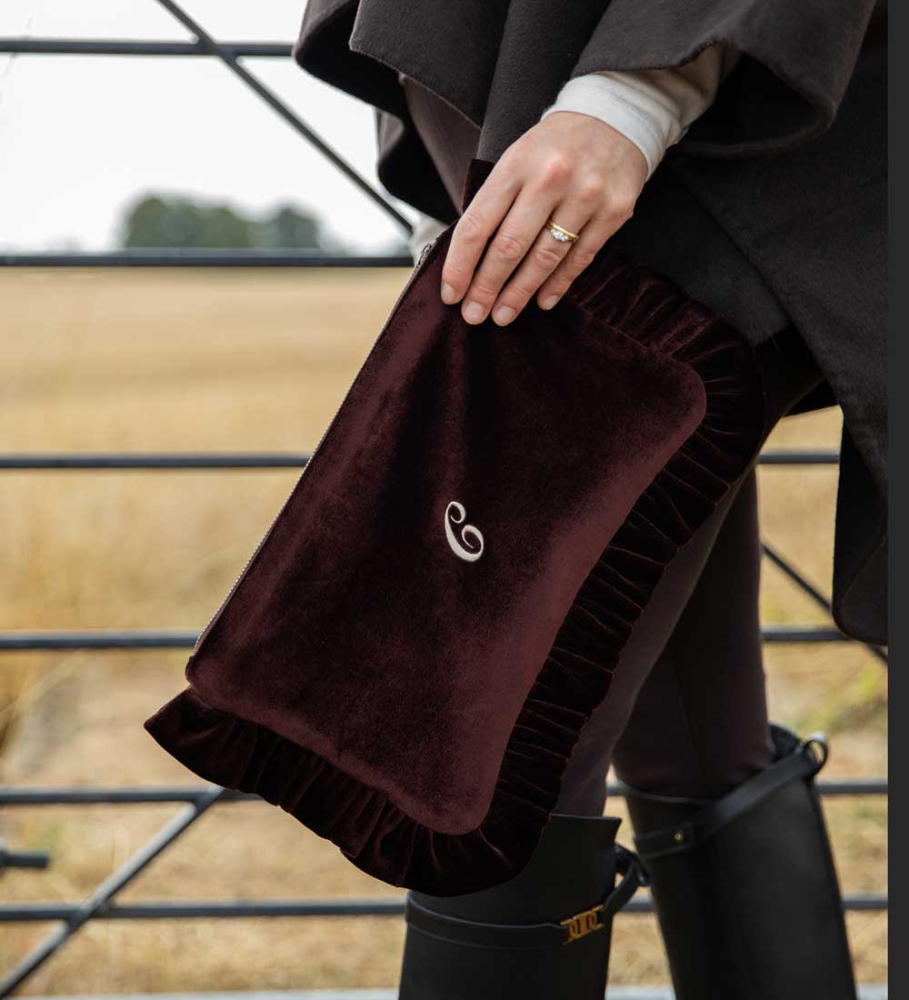 A person outdoors holds a plush brown velvet pouch with a small white embroidered swirl logo. The scene includes a field in the background and black rain boots.