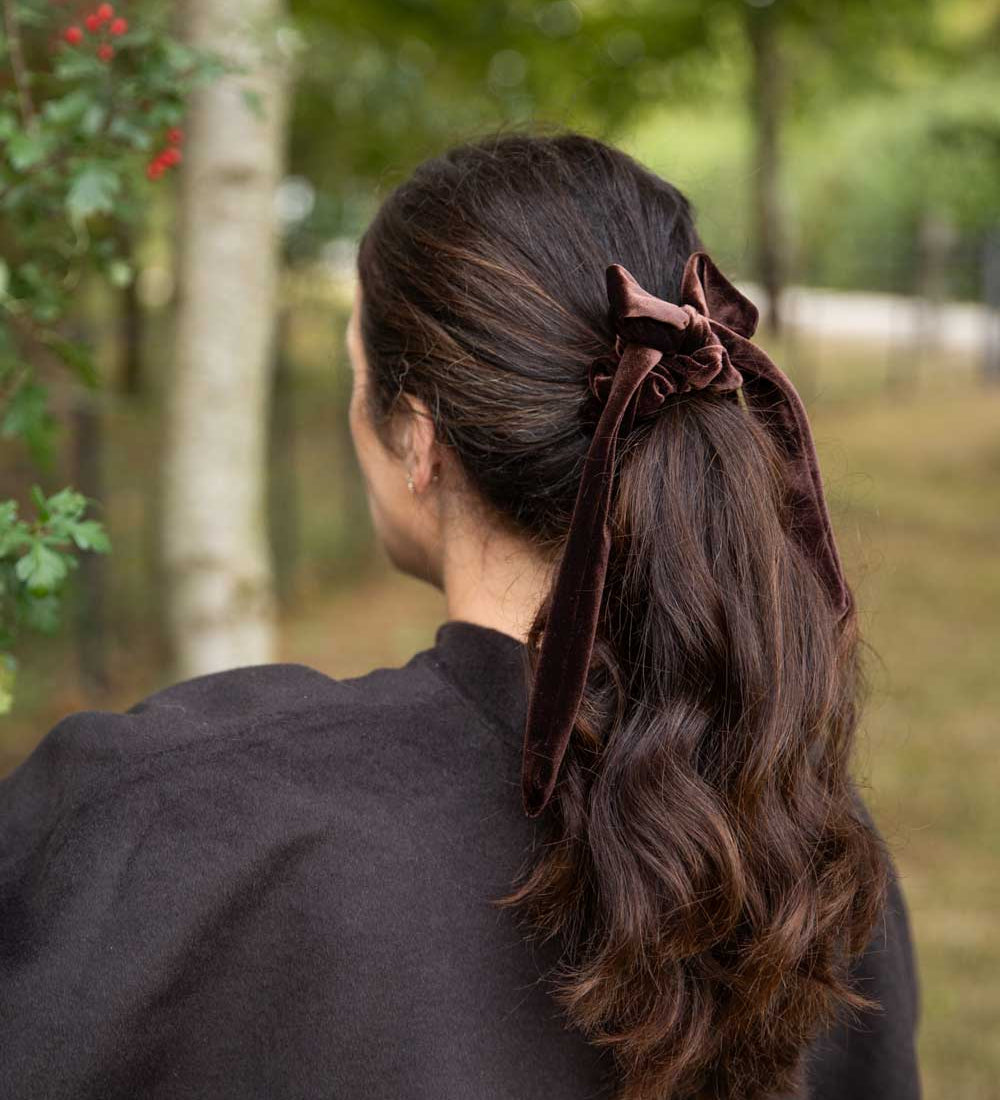 A woman viewed from behind outdoors, wearing a dark brown velvet hair tie or scrunchie that holds her long, wavy brown hair in a low ponytail.