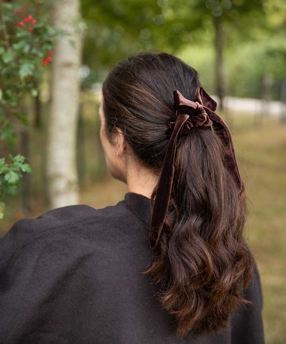 A woman viewed from behind outdoors, wearing a dark brown velvet hair tie or scrunchie that holds her long, wavy brown hair in a low ponytail.
