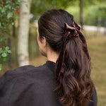 A woman viewed from behind outdoors, wearing a dark brown velvet hair tie or scrunchie that holds her long, wavy brown hair in a low ponytail.