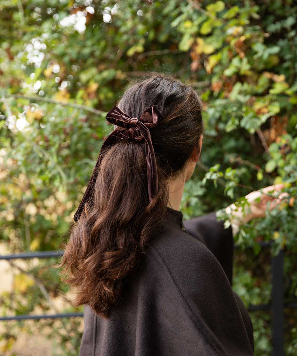 A woman viewed from behind wearing a dark brown velvet bow hair accessory tied to her ponytail, set against a blurred background of green foliage.