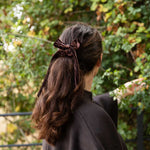 A woman viewed from behind wearing a dark brown velvet bow hair accessory tied to her ponytail, set against a blurred background of green foliage.