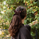 A woman viewed from behind wearing a dark brown velvet bow hair accessory tied to her ponytail, set against a blurred background of green foliage.