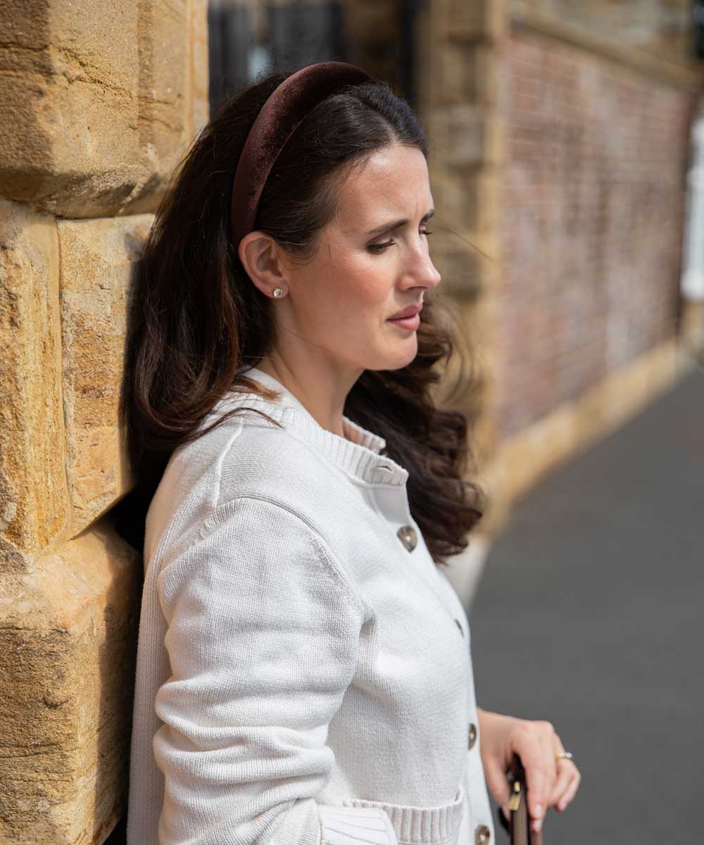 A woman in a white knit cardigan and a brown velvet headband leans against a stone wall outdoors.