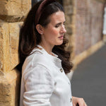 A woman in a white knit cardigan and a brown velvet headband leans against a stone wall outdoors.