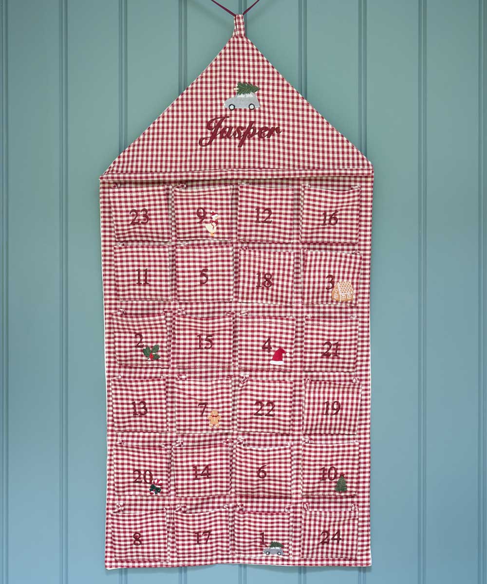 A red and white gingham house-shaped Advent calendar hanging on a light blue paneled wall. The top section is personalized with the name 'Jasper' in red script, and the 24 individual pockets are numbered in burgundy.