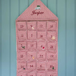 A red and white gingham house-shaped Advent calendar hanging on a light blue paneled wall. The top section is personalized with the name 'Jasper' in red script, and the 24 individual pockets are numbered in burgundy.
