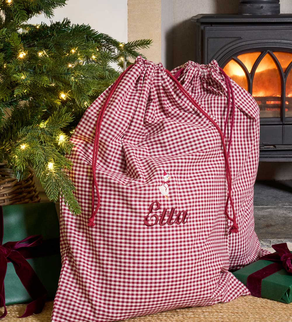 A personalized Christmas gift bag in a red and white gingham check pattern with red drawstrings and the name 'Ellen' embroidered on the front. The bag is styled in a cozy holiday setting next to a lit Christmas tree and a wood-burning stove.