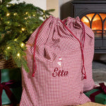 A personalized Christmas gift bag in a red and white gingham check pattern with red drawstrings and the name 'Ellen' embroidered on the front. The bag is styled in a cozy holiday setting next to a lit Christmas tree and a wood-burning stove.