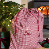 A personalized Christmas gift bag in a red and white gingham check pattern with red drawstrings and the name 'Ellen' embroidered on the front. The bag is styled in a cozy holiday setting next to a lit Christmas tree and a wood-burning stove.