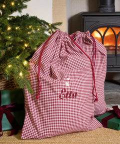 A personalized Christmas gift bag in a red and white gingham check pattern with red drawstrings and the name 'Ellen' embroidered on the front. The bag is styled in a cozy holiday setting next to a lit Christmas tree and a wood-burning stove.