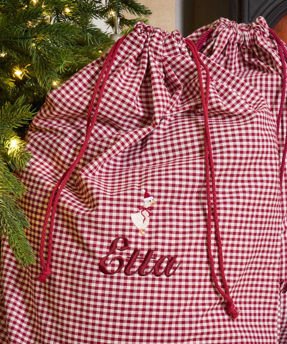 A burgundy and white gingham Christmas-themed storage bag with red drawstrings, featuring a snowman graphic and the embroidered name 'Ella' in red script. The bag is positioned next to a decorated Christmas tree with warm lights.