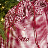 A burgundy and white gingham Christmas-themed storage bag with red drawstrings, featuring a snowman graphic and the embroidered name 'Ella' in red script. The bag is positioned next to a decorated Christmas tree with warm lights.