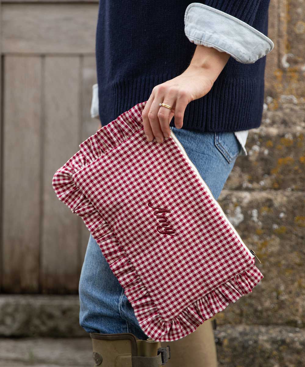 A person in a navy sweater and blue jeans holds a red and white gingham clutch bag with a ruffled edge and embroidered text, set against a rustic stone wall background.