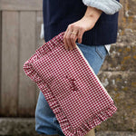 A person in a navy sweater and blue jeans holds a red and white gingham clutch bag with a ruffled edge and embroidered text, set against a rustic stone wall background.