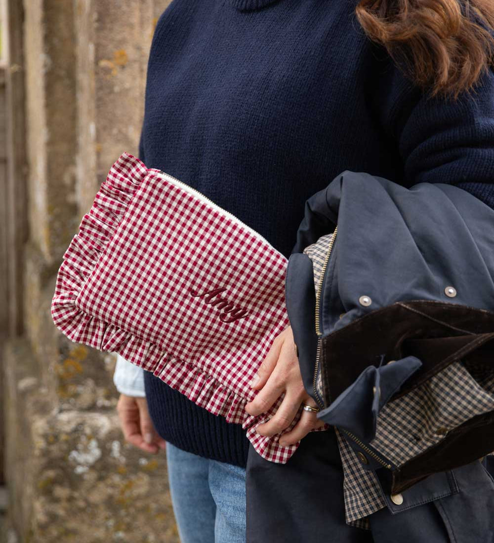 A close-up lifestyle shot of a person holding a red and white gingham clutch bag with 'Lily' embroidered on it, alongside a navy blue jacket.