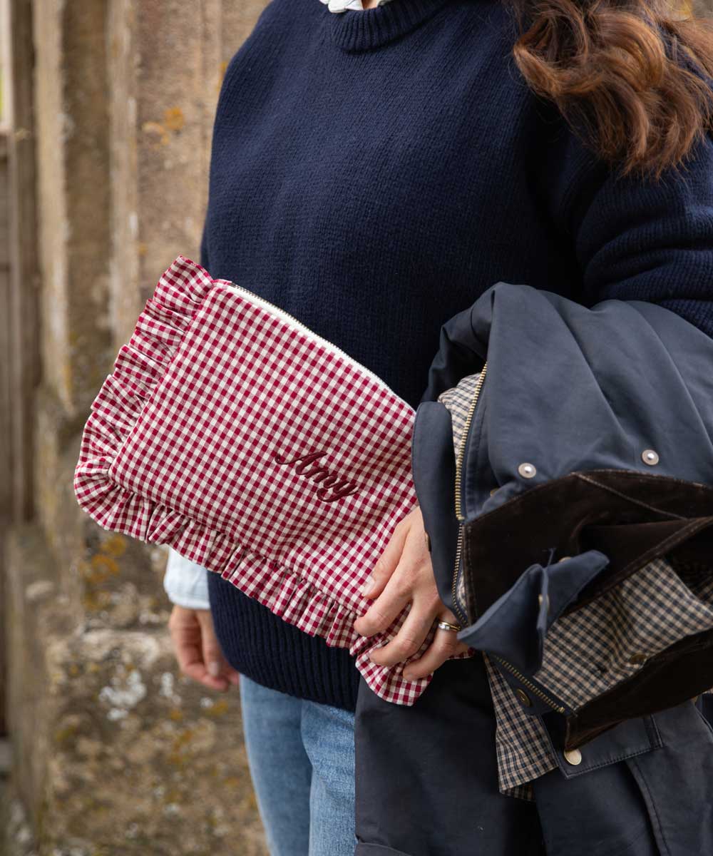 A close-up lifestyle shot of a person holding a red and white gingham clutch bag with 'Lily' embroidered on it, alongside a navy blue jacket.