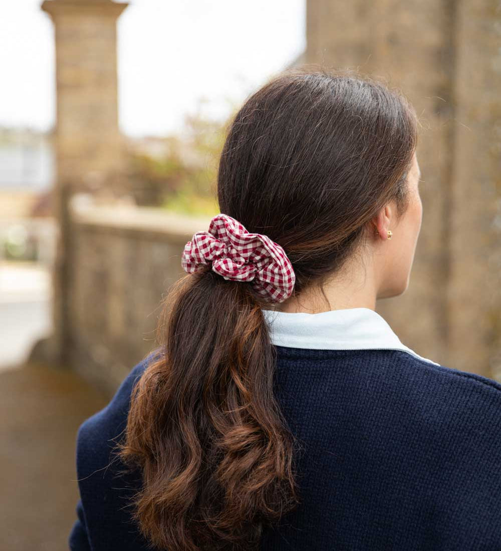 A woman viewed from behind wearing a red and white gingham check scrunchie to hold her dark brown hair in a ponytail, wearing a navy blue sweater over a white collared shirt, with a blurred stone bridge background.