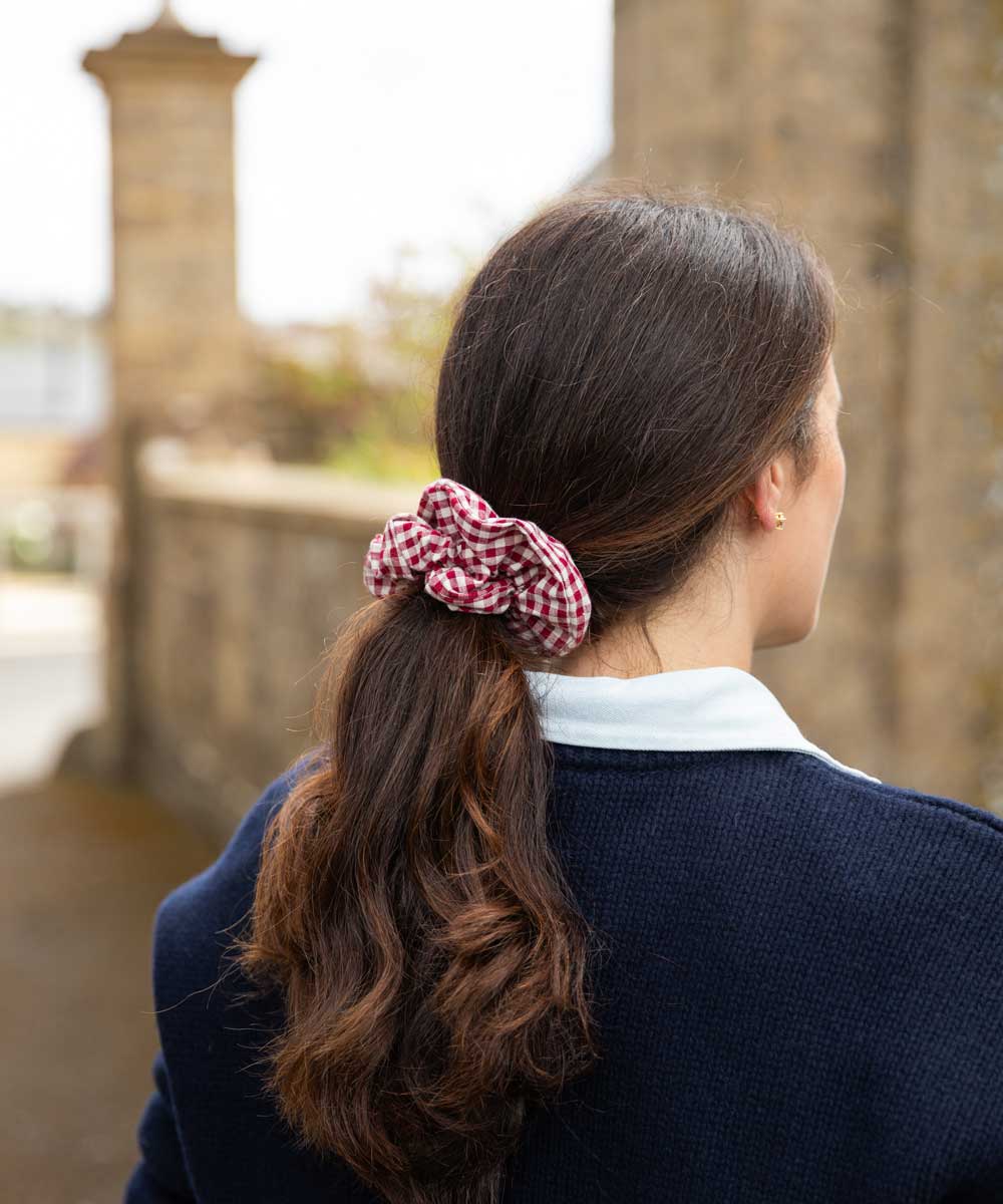 A woman viewed from behind wearing a red and white gingham check scrunchie to hold her dark brown hair in a ponytail, wearing a navy blue sweater over a white collared shirt, with a blurred stone bridge background.