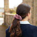 A woman viewed from behind wearing a red and white gingham check scrunchie to hold her dark brown hair in a ponytail, wearing a navy blue sweater over a white collared shirt, with a blurred stone bridge background.