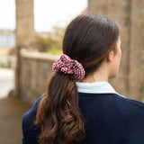 A woman viewed from behind wearing a red and white gingham check scrunchie to hold her dark brown hair in a ponytail, wearing a navy blue sweater over a white collared shirt, with a blurred stone bridge background.