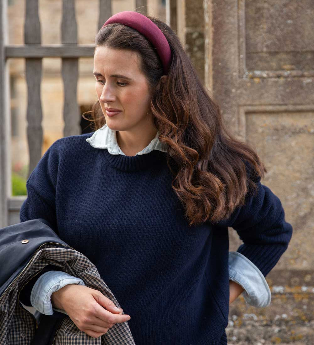 A woman wearing a navy blue knit sweater over a light blue collared shirt, styled with a burgundy linen headband and holding a dark jacket, set against a stone wall background.