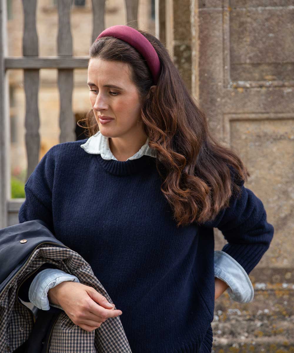 A woman wearing a navy blue knit sweater over a light blue collared shirt, styled with a burgundy linen headband and holding a dark jacket, set against a stone wall background.