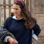 A woman wearing a navy blue knit sweater over a light blue collared shirt, styled with a burgundy linen headband and holding a dark jacket, set against a stone wall background.