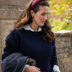 A woman wearing a navy blue knit sweater over a white collared blouse, with a burgundy headband and holding a dark coat, set against a blurred stone wall and garden background.