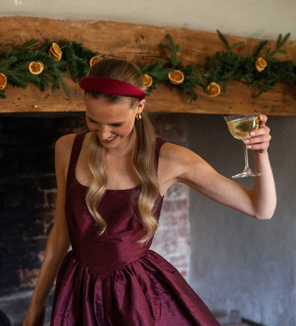 Woman in a burgundy dress and burgundy satin headband holding a glass of white wine in front of a decorated fireplace.