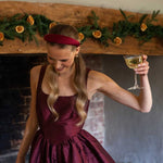 Woman in a burgundy dress and burgundy satin headband holding a glass of white wine in front of a decorated fireplace.