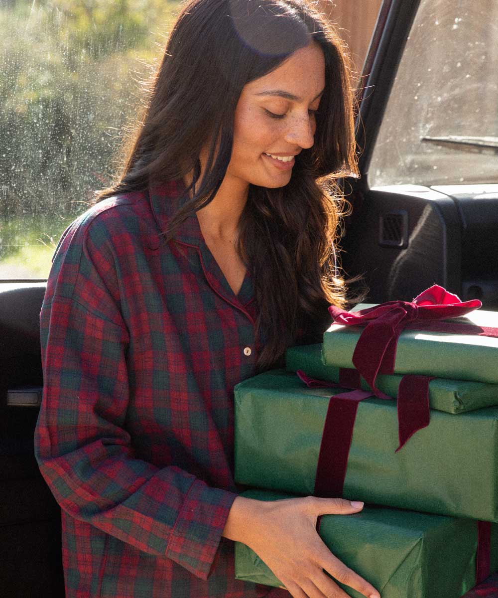 A woman in burgundy and green plaid pyjamas smiles while carrying three wrapped gift boxes with green paper and dark red ribbons inside a vehicle.