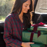 A woman in burgundy and green plaid pyjamas smiles while carrying three wrapped gift boxes with green paper and dark red ribbons inside a vehicle.