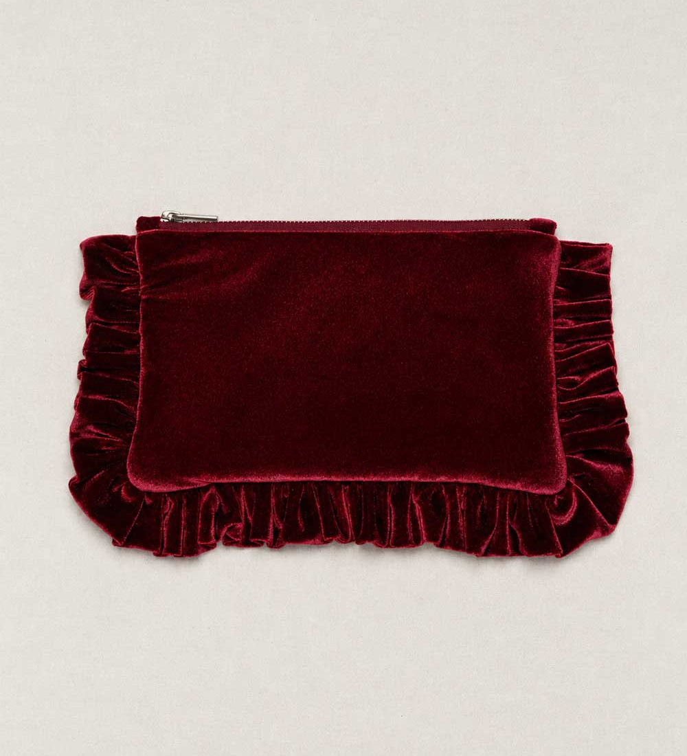 A deep burgundy velvet clutch pouch with a top zipper and decorative ruffled trim on the sides and bottom, displayed flat against a light beige background.