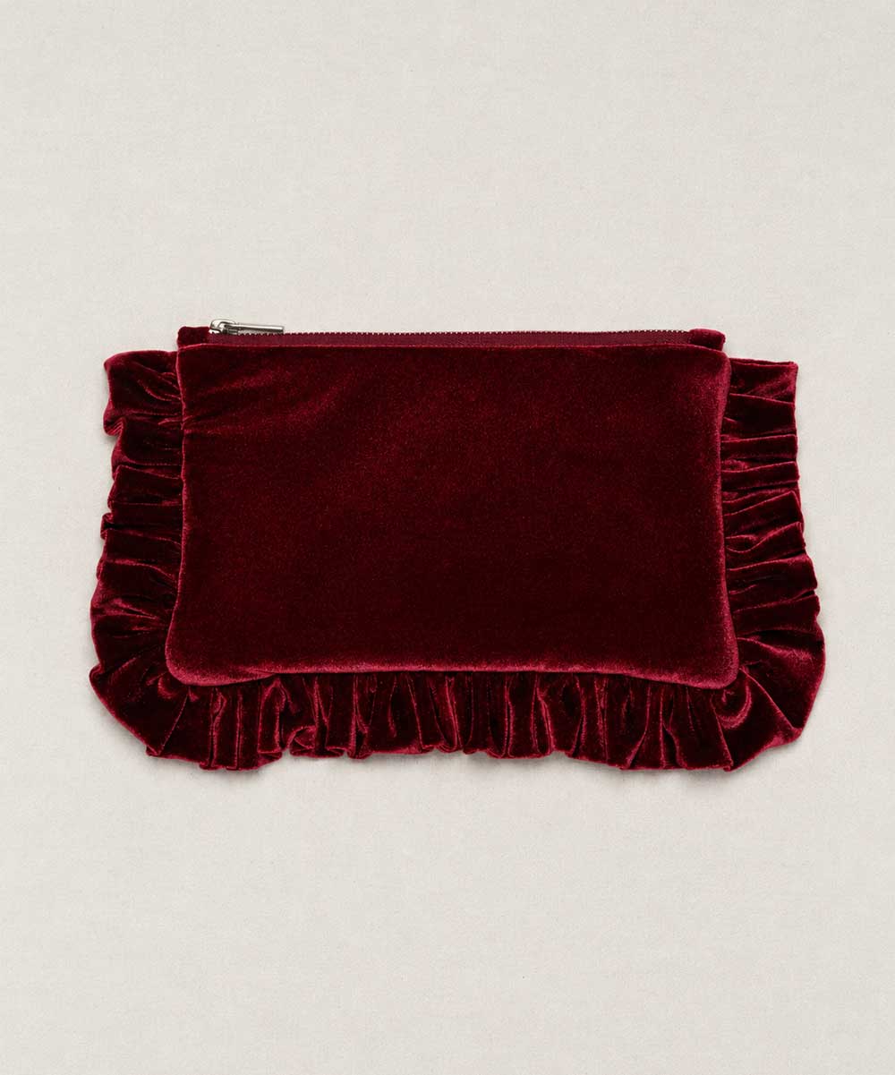 A deep burgundy velvet clutch pouch with a top zipper and decorative ruffled trim on the sides and bottom, displayed flat against a light beige background.