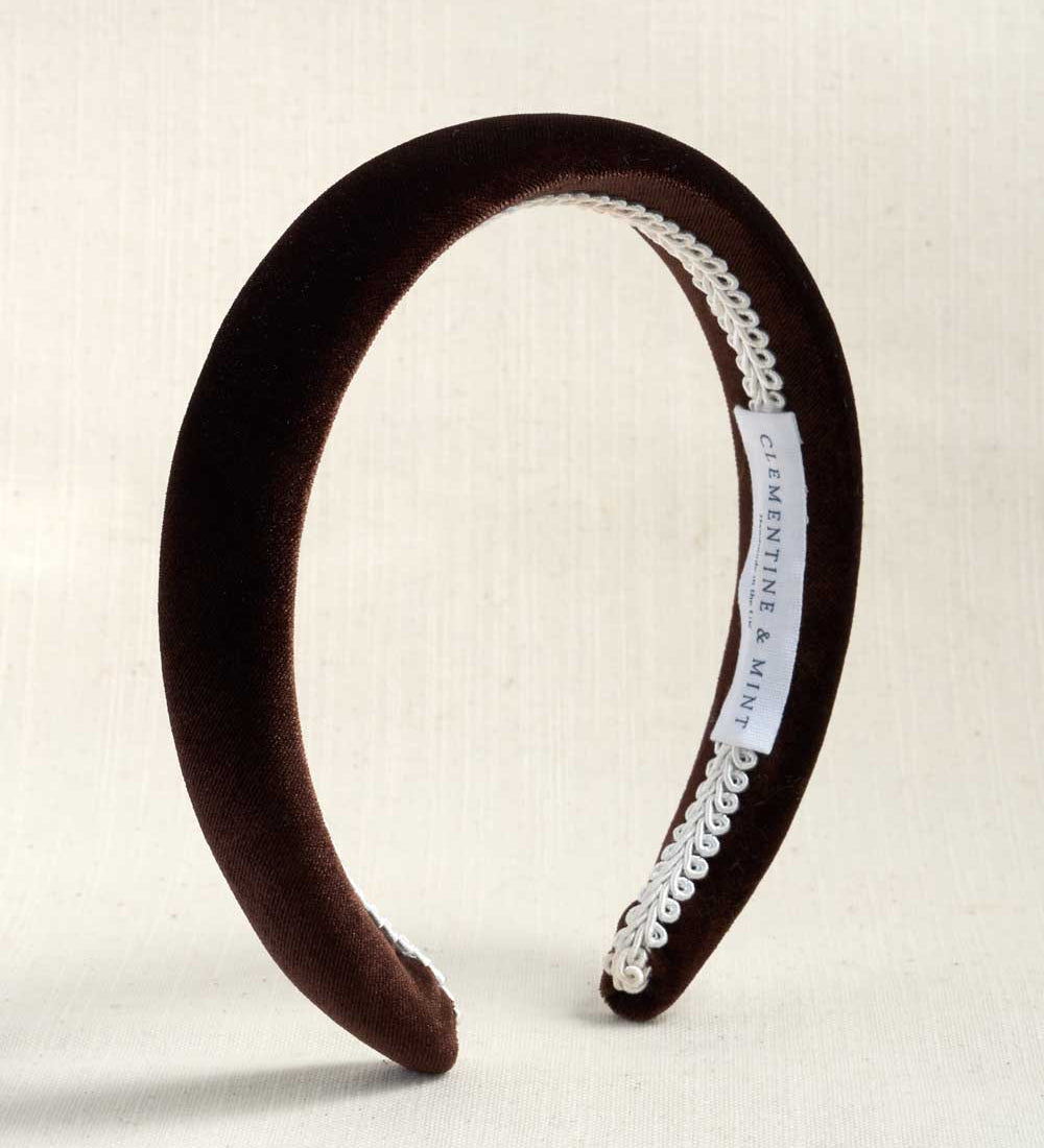 A dark brown velvet headband with a white lace trim and a white brand label, displayed on a light beige textured background.
